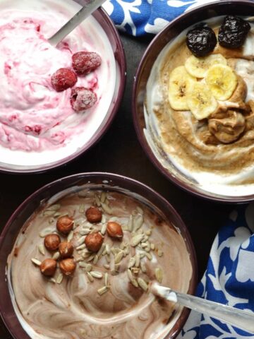 Flavoured mocha, raspberry, prune yogurt in purple bowls with spoons and blue-and-white clothe on dark surface.