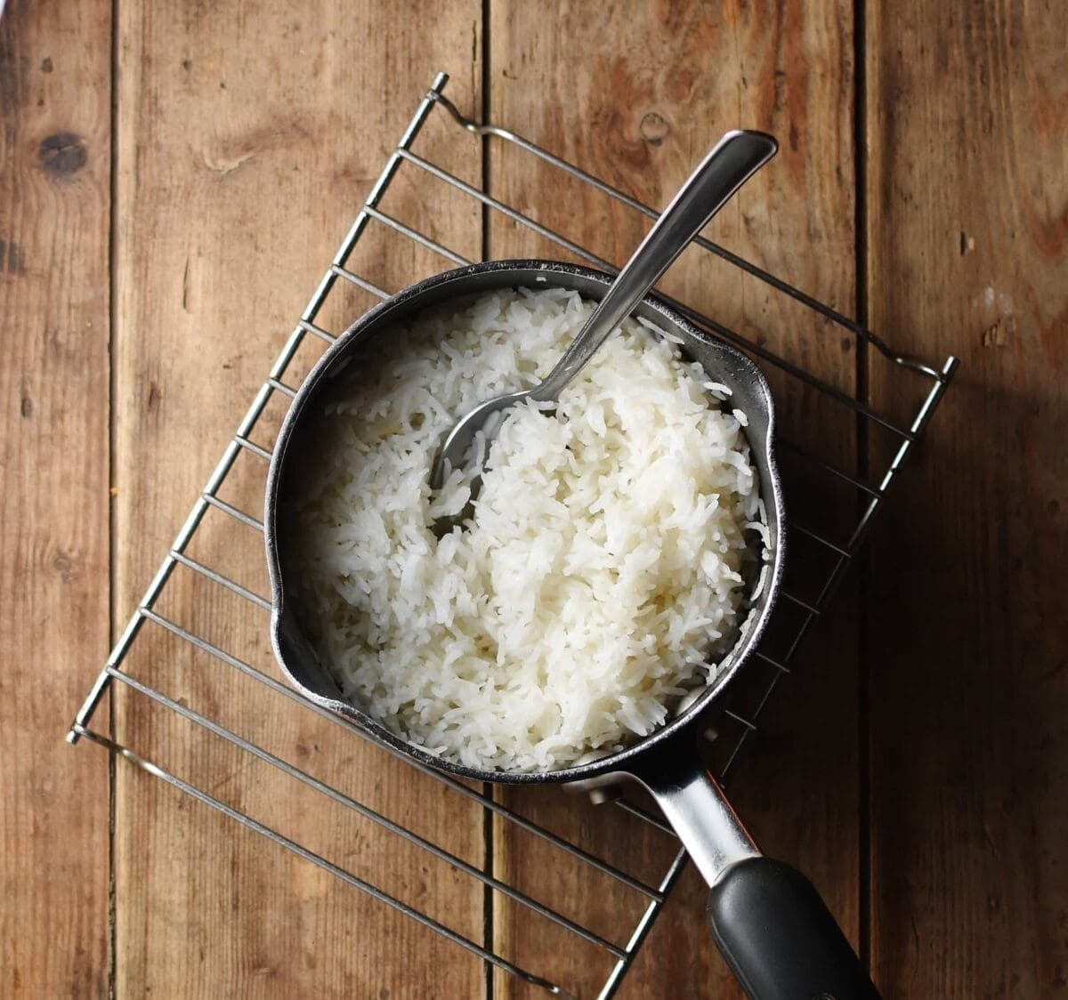 Cooked rice in saucepan with spoon on top of cooling rack.