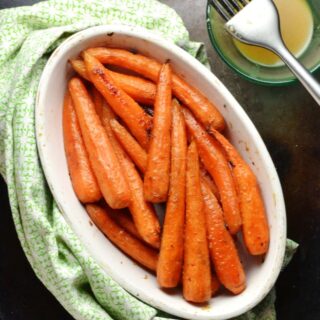 Honey roasted carrots in white oval dish wrapped in green cloth and small dish with dressing and baking brush in top right corner.