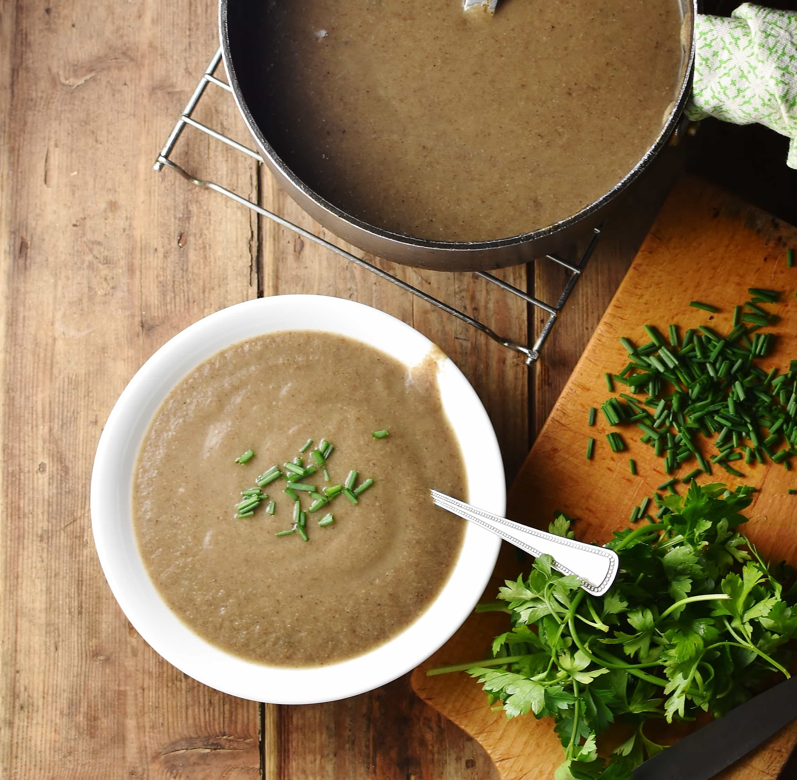 Top down view of creamy mushroom soup in white bowl with spoon, fresh herbs on cutting board to the right and soup in pot at the top.