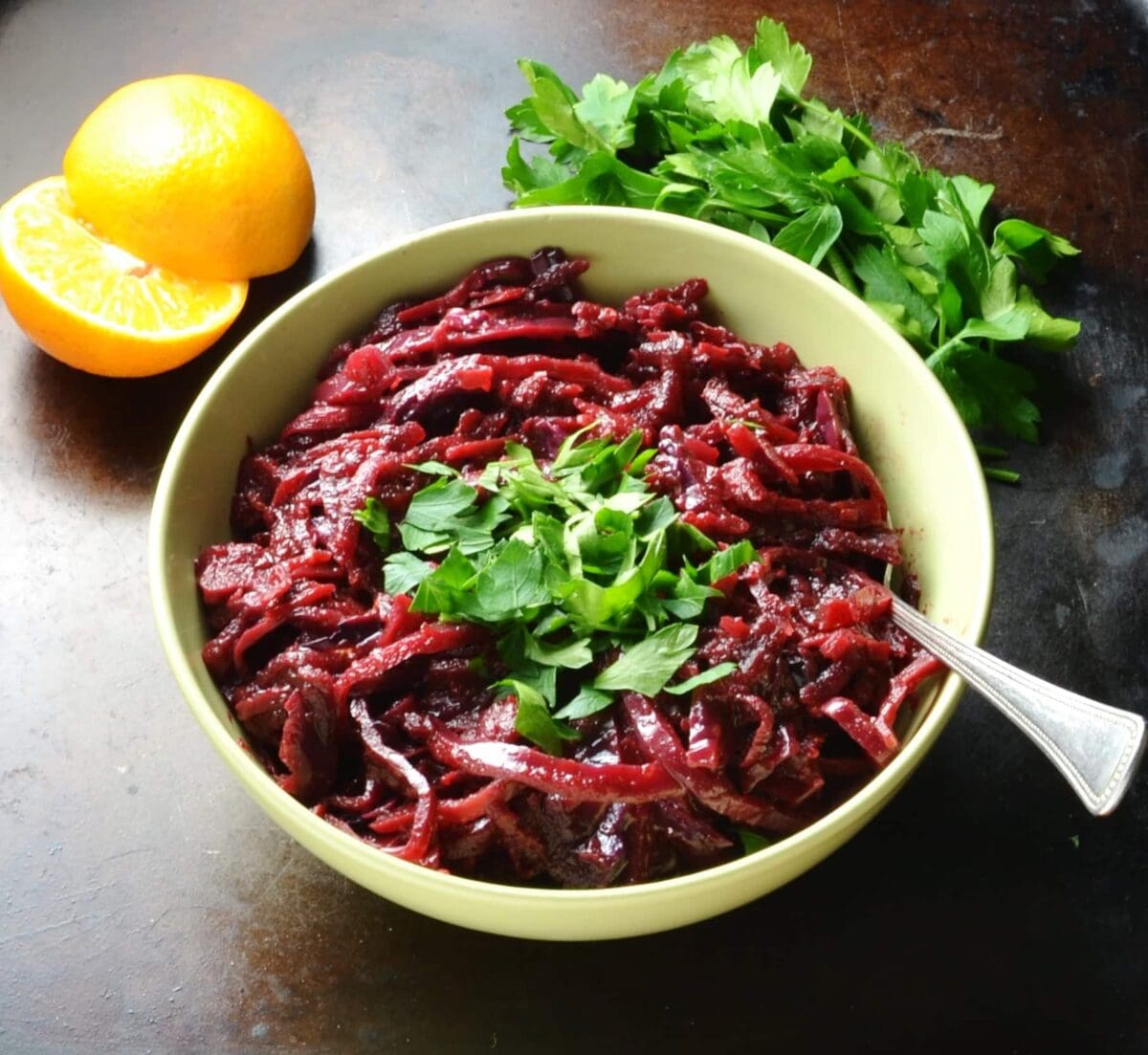 Braised red cabbage in green bowl with spoon and clementine and herbs in background.