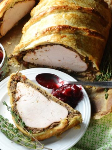 Turkey wellington on white plate with rosemary, spoon and cranberry sauce.