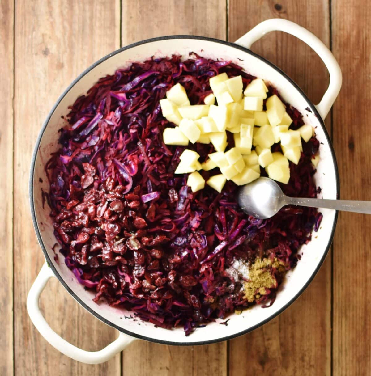 Shredded red cabbage, spices and cubed apple in large white pan with spoon.