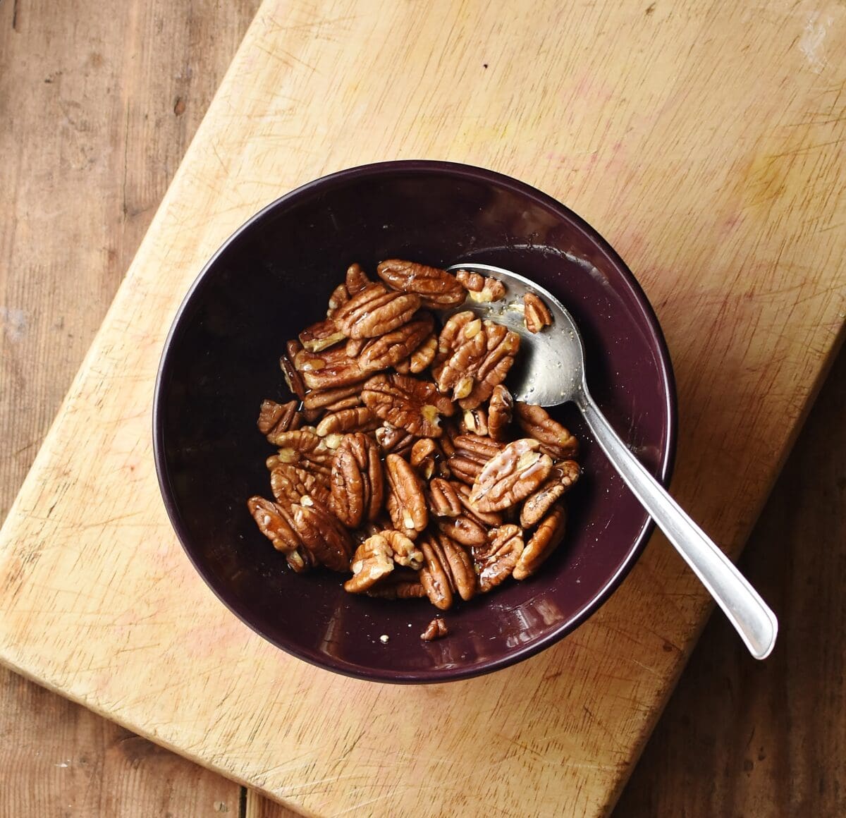Pecans with maple glaze in purple bowl with spoon on top of wooden board.