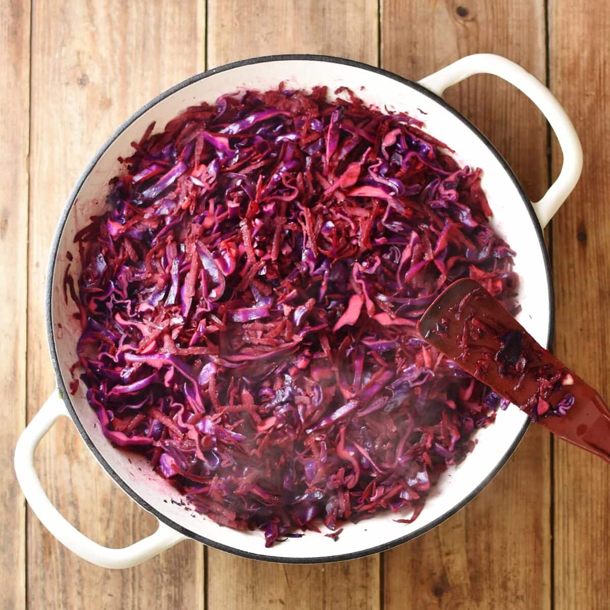 Braise shredded red cabbage with red spatula in large white pan.