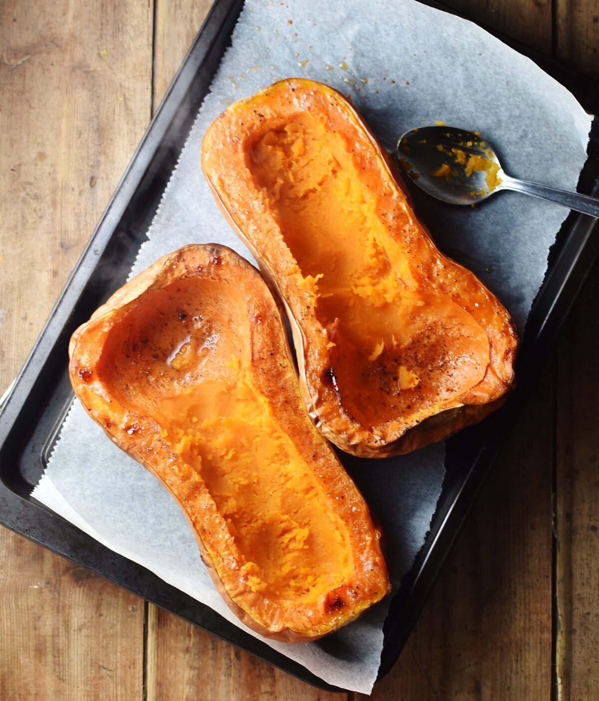 Roasted butternut squash halves with scooped out middle on top of baking sheet lined with paper and spoon.