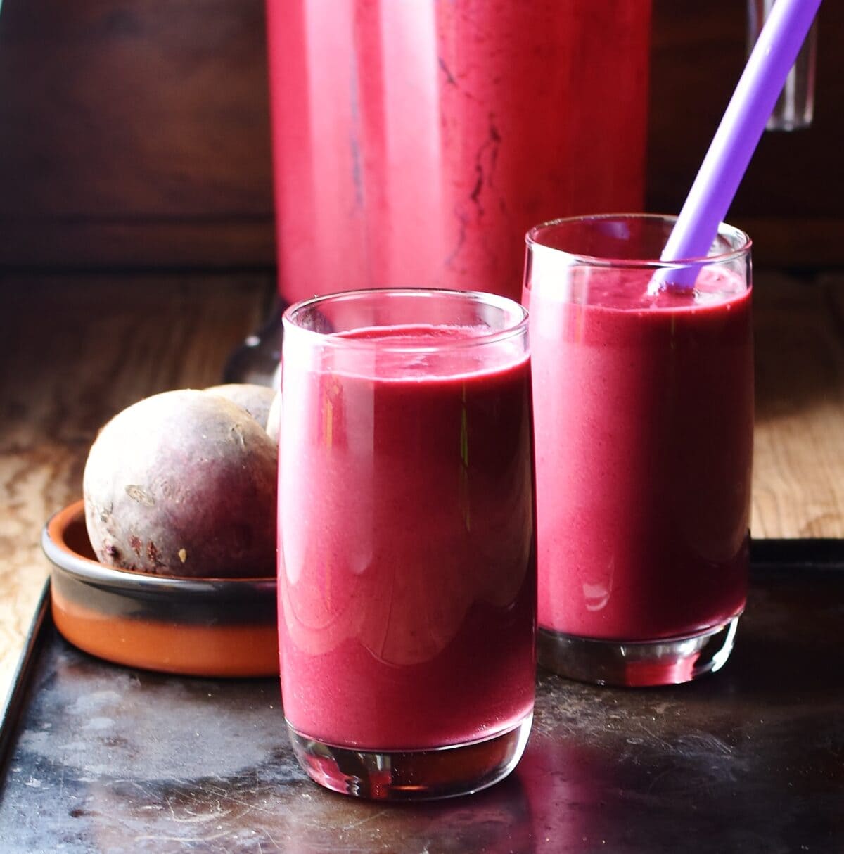 Side view of beet smoothie in 2 glasses and blender and beetroot in background.