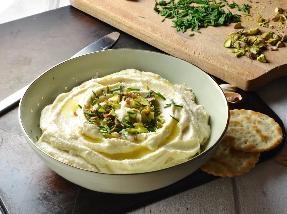 Close-up image of cauliflower dip garnished with chives and pistachios in cream colour bowl.