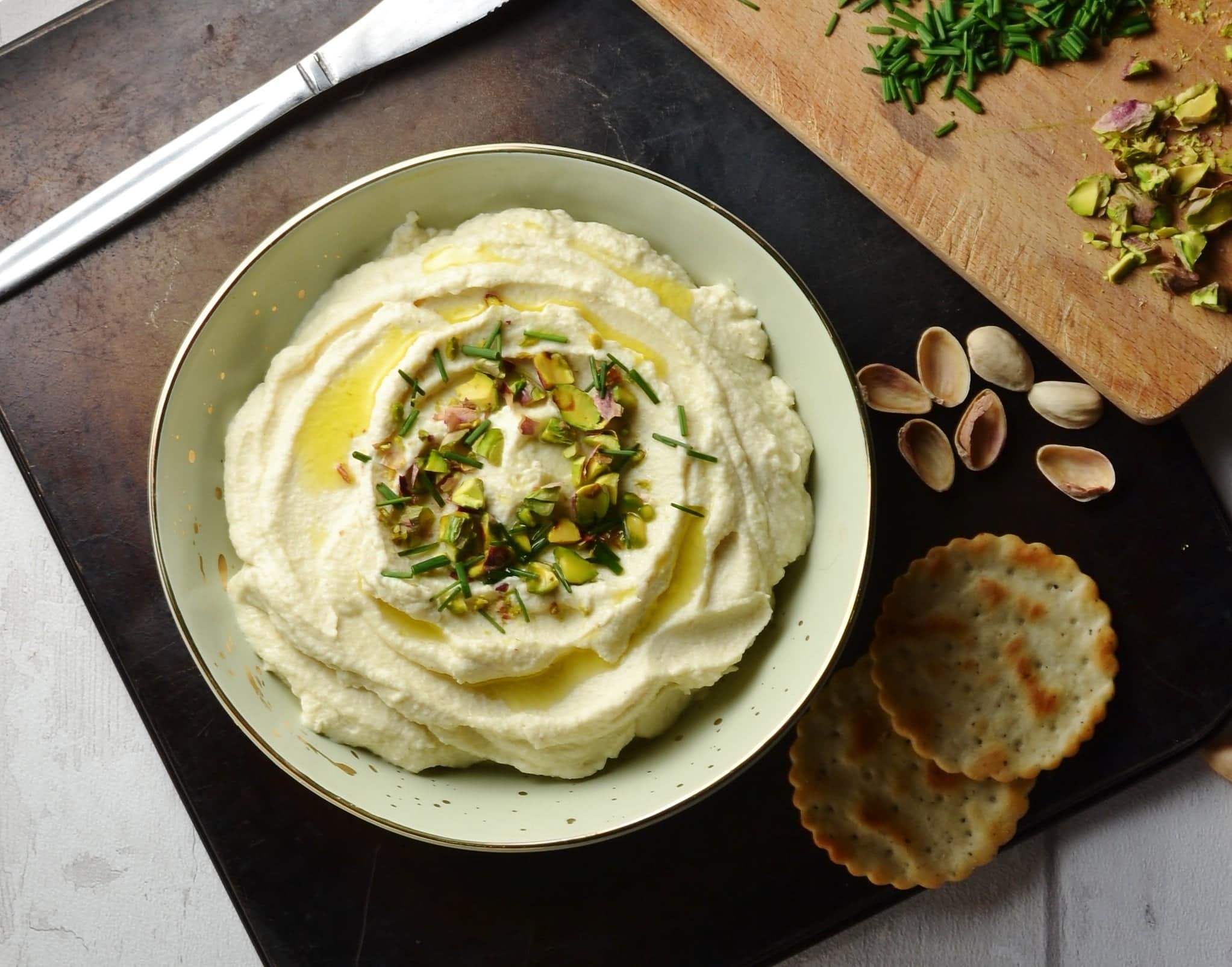 Top down view of cauliflower dip in cream bowl on top of tray with crackers, nuts, herbs and knife in background.. 
