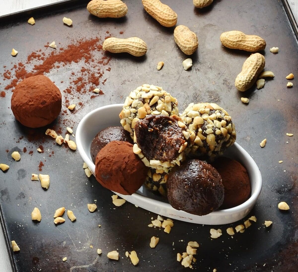 Close-up view of nut and cocoa coated energy balls in white dish with peanuts in background on metallic tray.