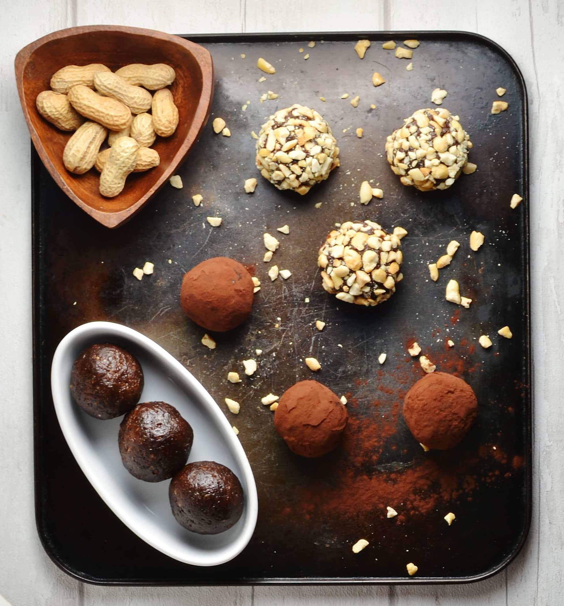 Top down view of nut and cocoa coated energy balls in white dish with peanuts in brown dish on top of dark tray.