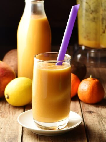 Sweet potato smoothie in glass with purple straw, lemon, tangerine, mango, smoothie in bottle and blender in background.