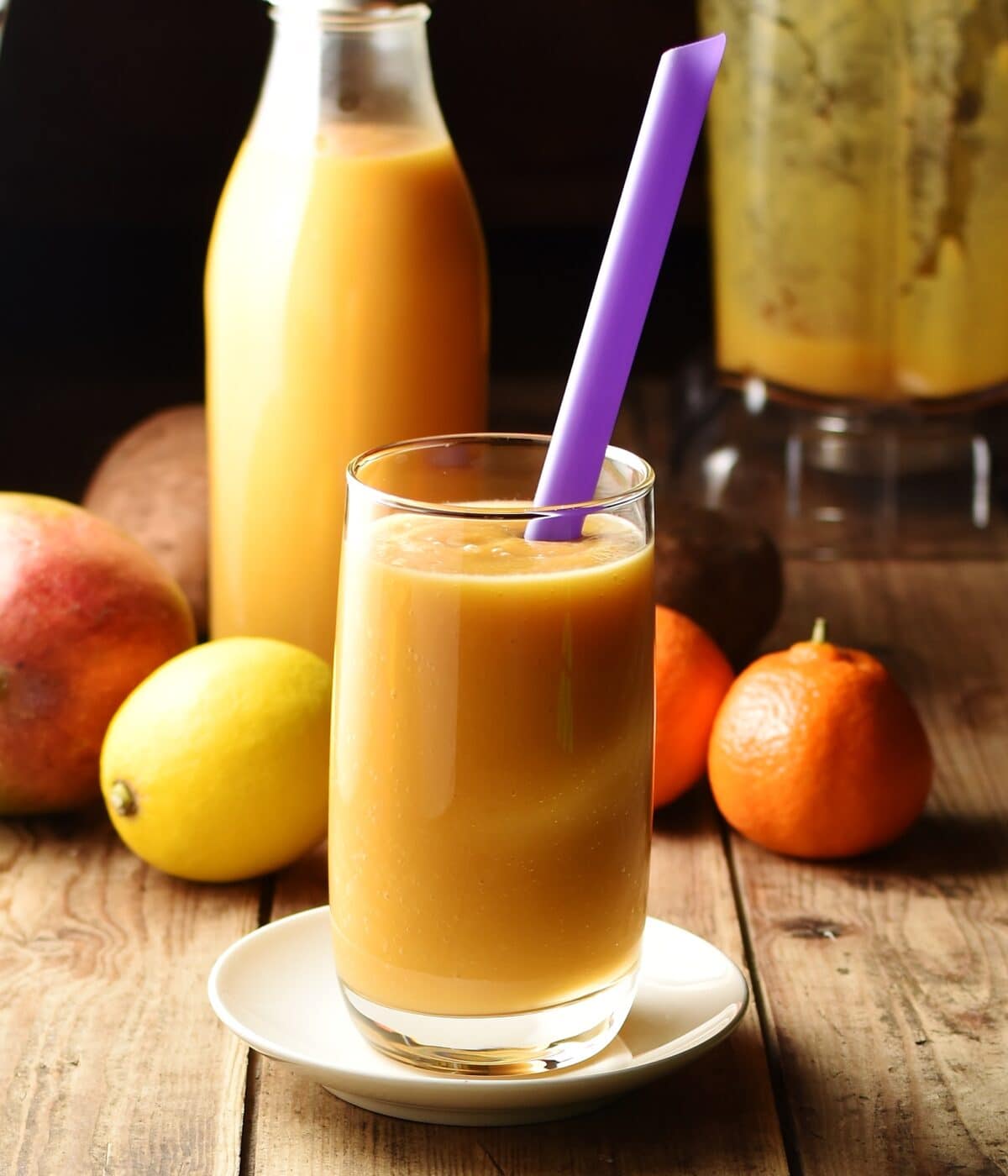Sweet potato smoothie in glass with purple straw, lemon, tangerine, mango, smoothie in bottle and blender in background.
