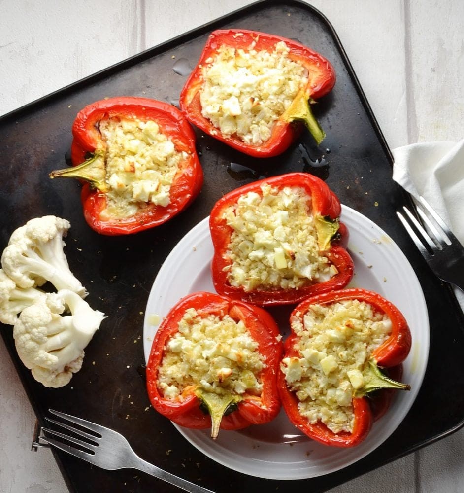 Cauliflower rice red stuffed peppers on white plate with cauliflower florets and forks on brown oven tray.