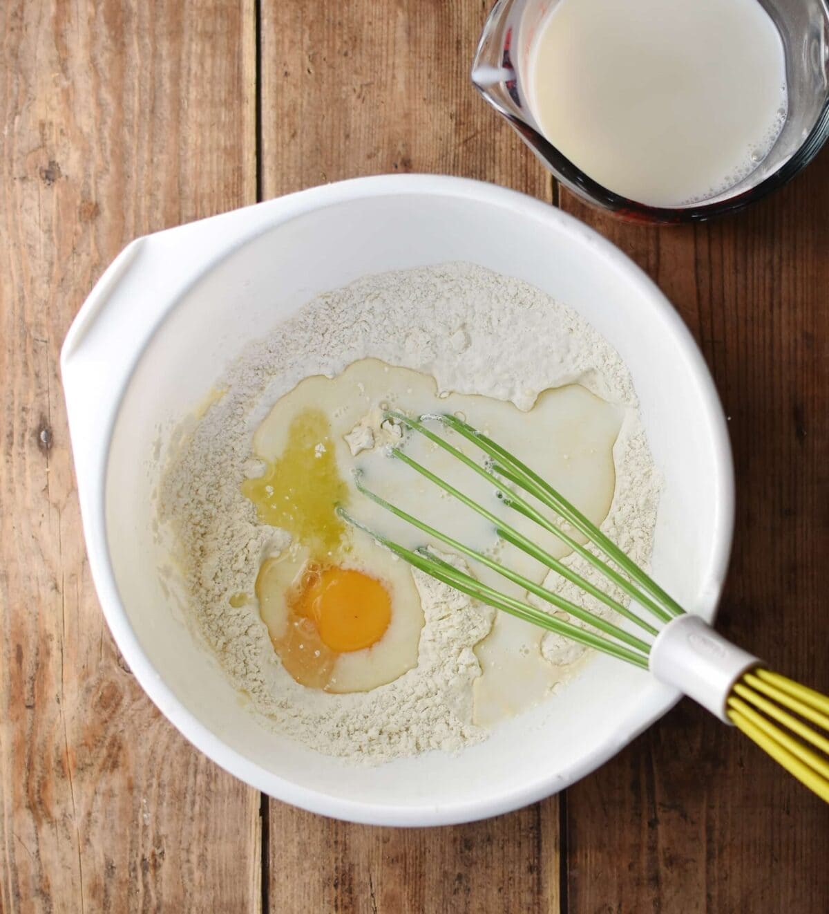Flour and egg inside large white bowl with green whisk and milk in measuring dish in top right corner.
