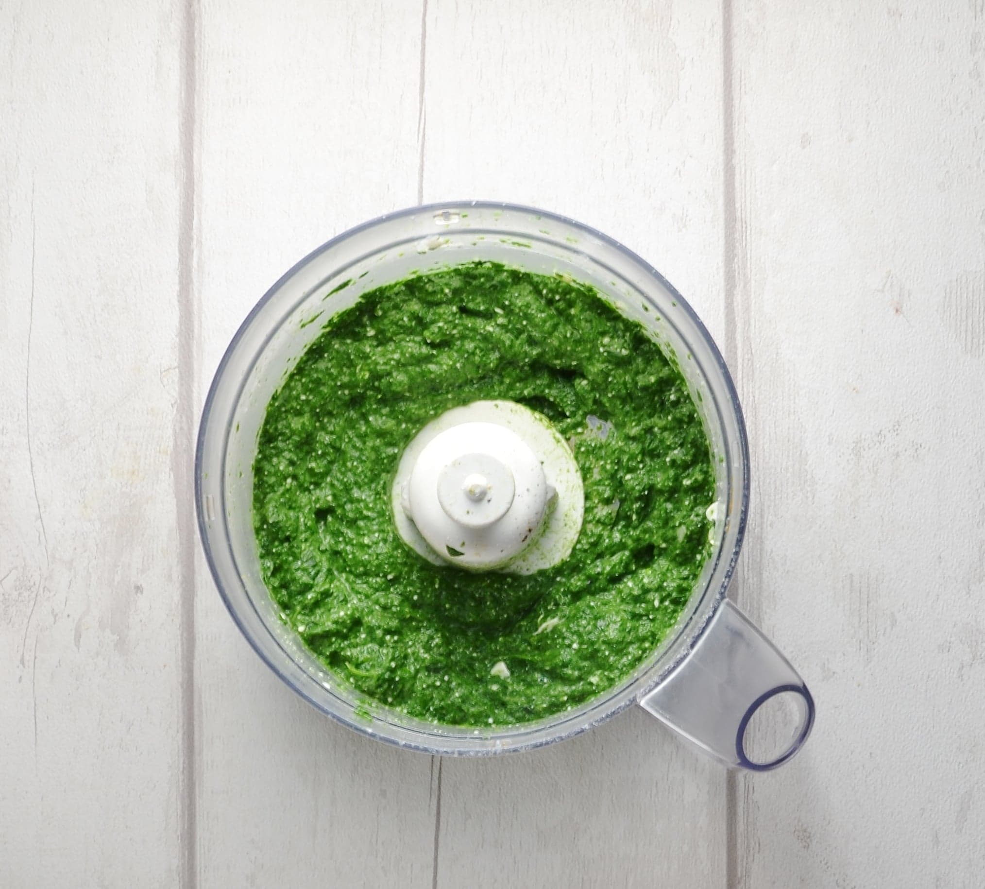 Top down view of spinach filling mixture in blender bowl on white wood background.
