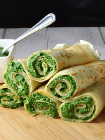 Crepes with spinach filling on light wooden board with white dish with spinach mixture and spoon in background.