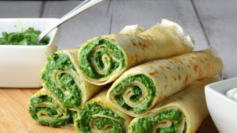 Crepes with spinach filling on light wooden board with white dish with spinach mixture and spoon in background.