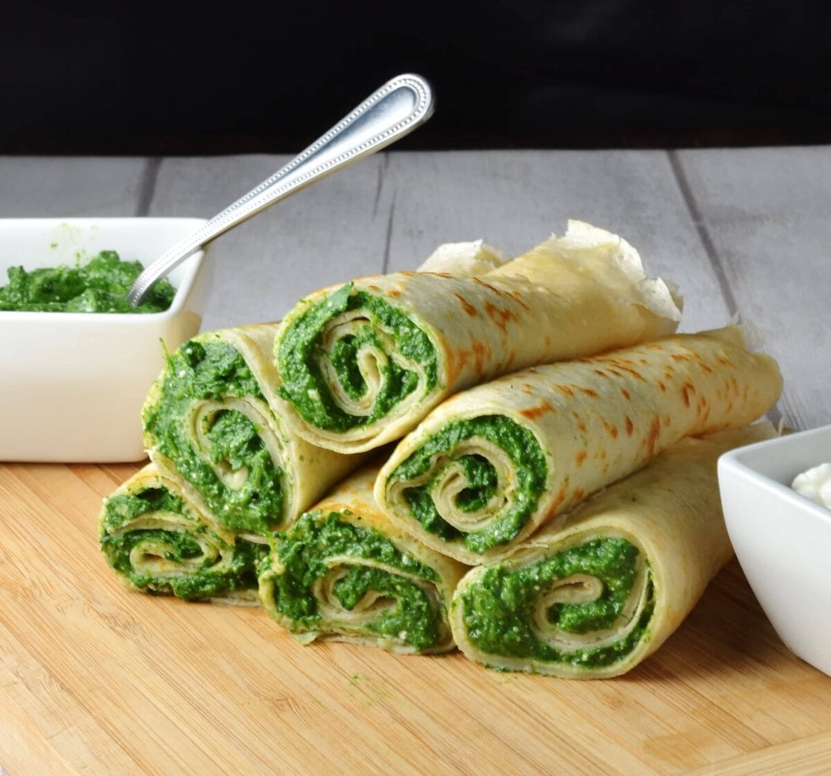 Crepes with spinach filling on light wooden board with white dish with spinach mixture and spoon in background.