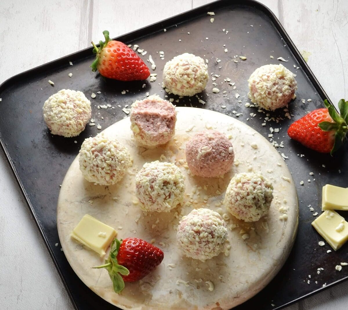 Strawberry cheesecake balls on round marble plate with strawberries and white chocolate on oven tray.