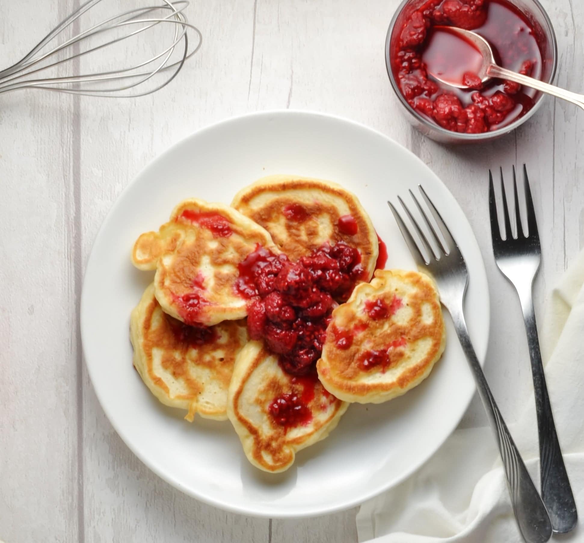 Polish pancakes with a dollop of raspberry compote on white plate with forks and raspberry compote in round dish and whisk in background.