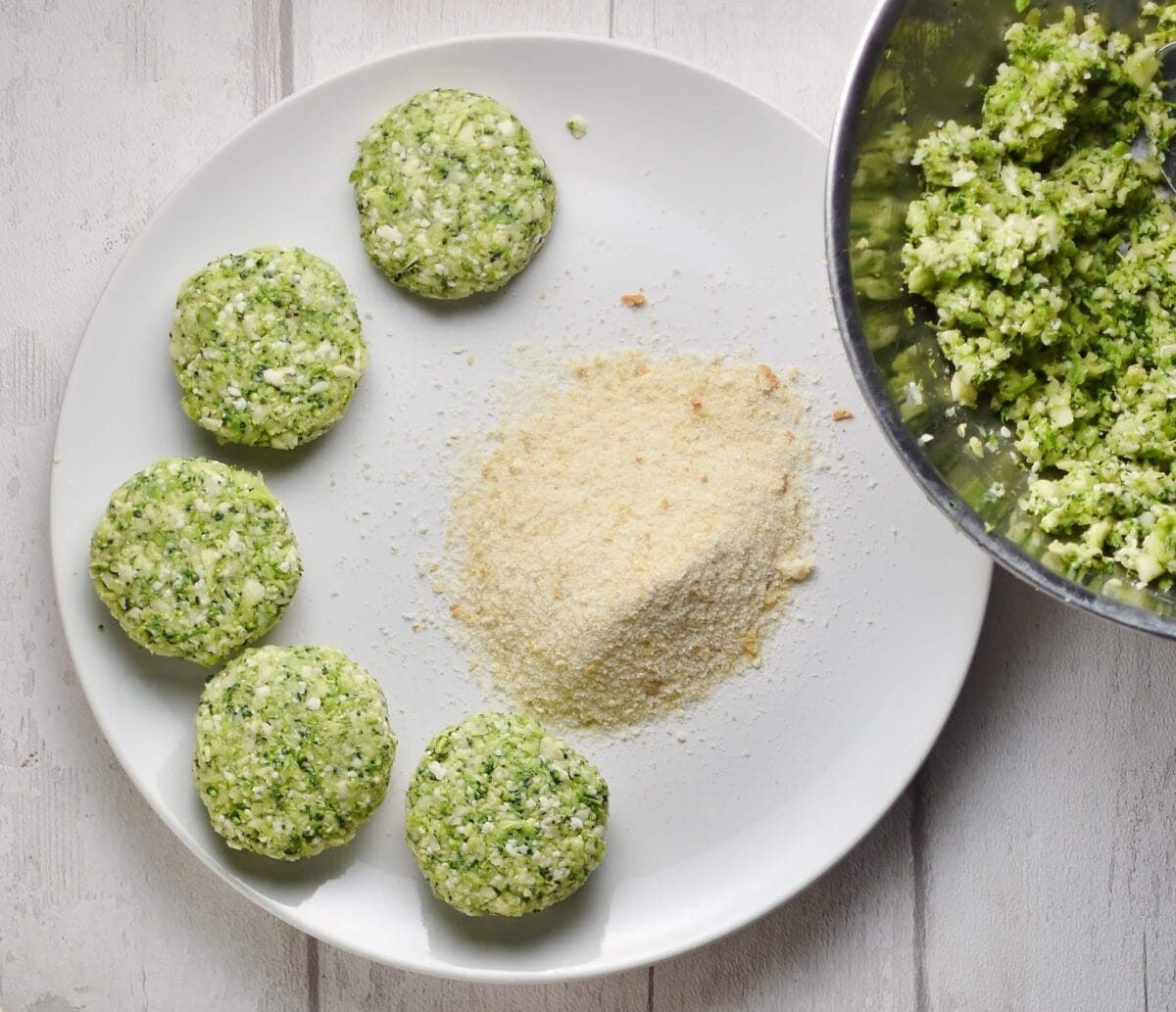 Top down view of raw broccoli patties with breadcrumbs on white plate.