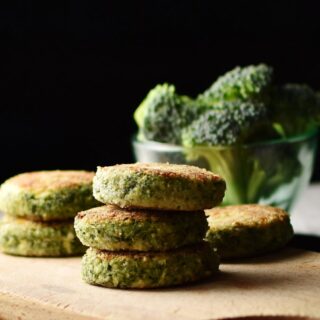 Side view of broccoli patties stacked on wooden board with broccoli florets in small dish in background.