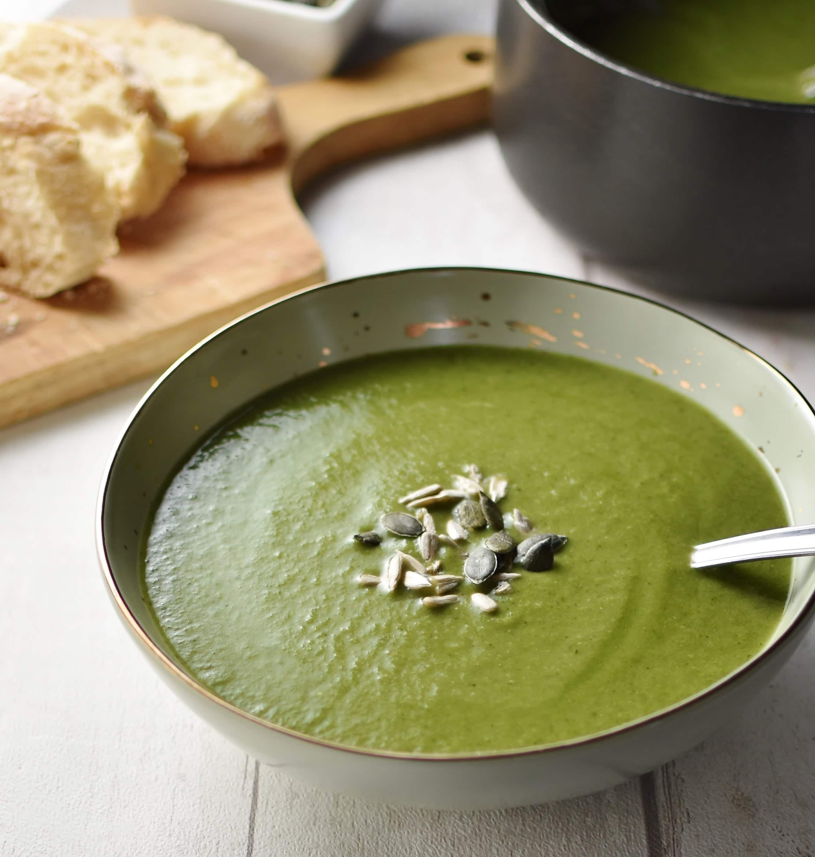 Side view of creamy broccoli spinach soup with seeds and spoon in green bowl, with black saucepan and slices of bread on top of cutting board in background.