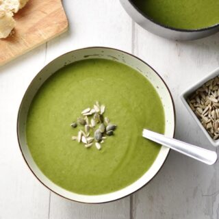 Top down view of broccoli spinach soup with seeds in grey bowl with spoon, seeds and saucepan with soup to right and wooden board in top left corner.