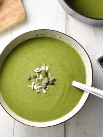 Top down view of broccoli spinach soup with seeds in grey bowl with spoon, seeds and saucepan with soup to right and wooden board in top left corner.