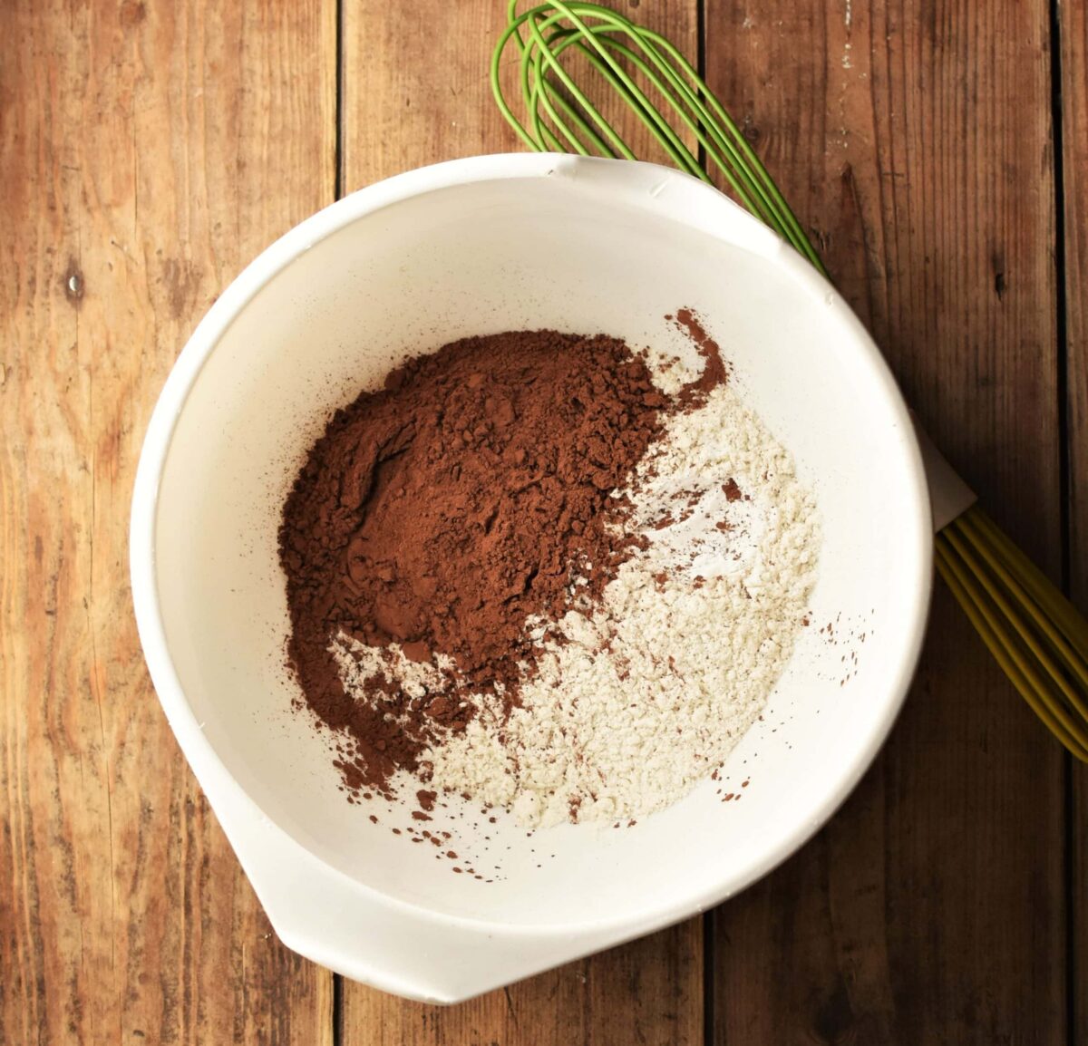 Flour and cocoa powder in large white bowl with green whisk.