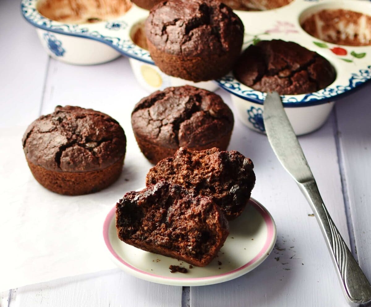 Side view of halved chocolate muffin on top of small white plate, with more muffins behind and inside ceramic muffin pan, with knife, in background.