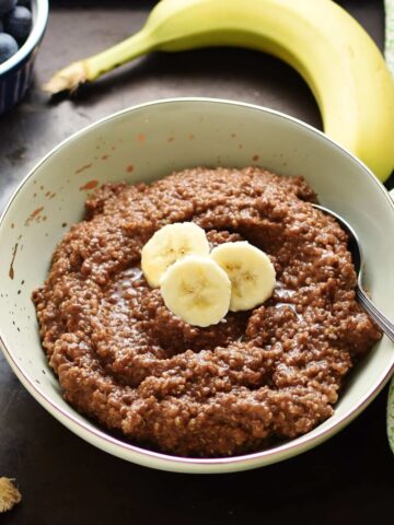 Quinoa chocolate porridge with slices of banana and spoon in light green bowl, with green cloth, banana and blueberries in small dish in background.