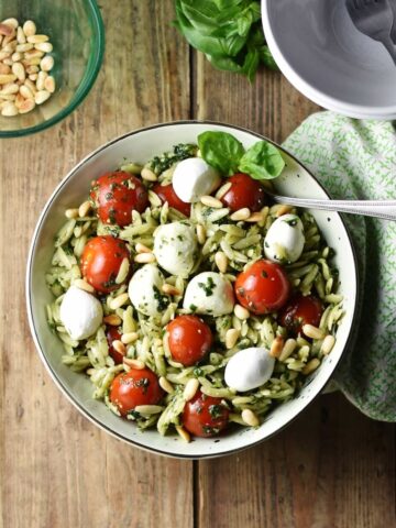 Top down view of orzo salad with pesto, cherry tomatoes, mini mozzarellas and spoon in white bowl wrapped in green cloth, with white bowl in top right and pine nuts in small dish in top left corner.