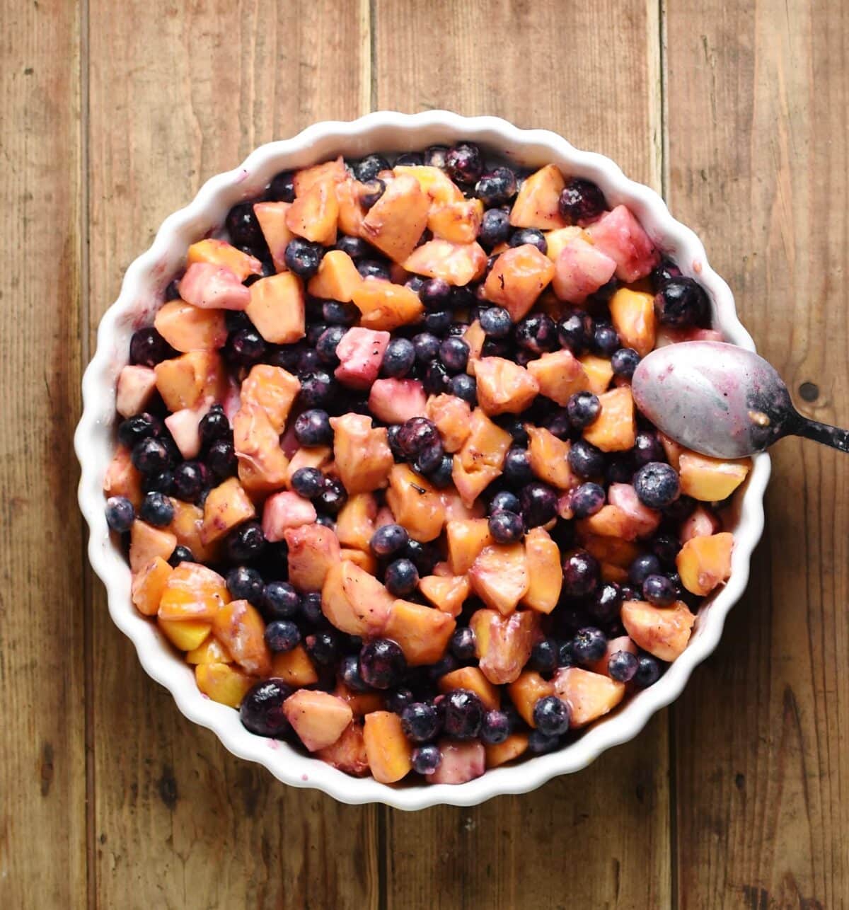 Top down view of cubed peaches and blueberries inside white round dish with spoon to the right.