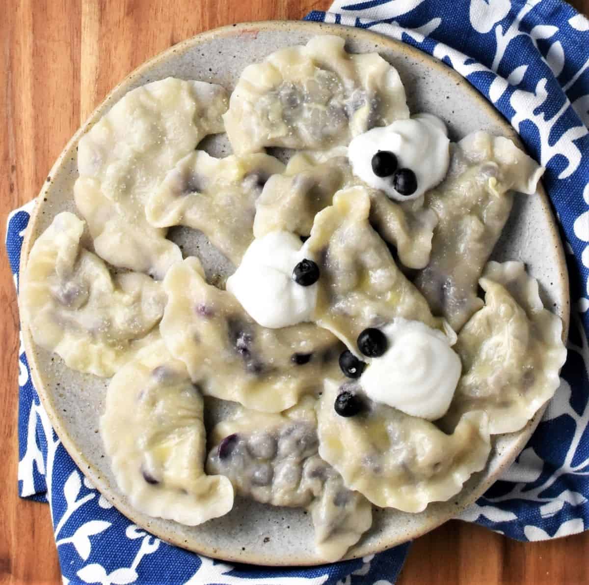 Blueberry pierogi with sour cream on top of plate with blue cloth in background.