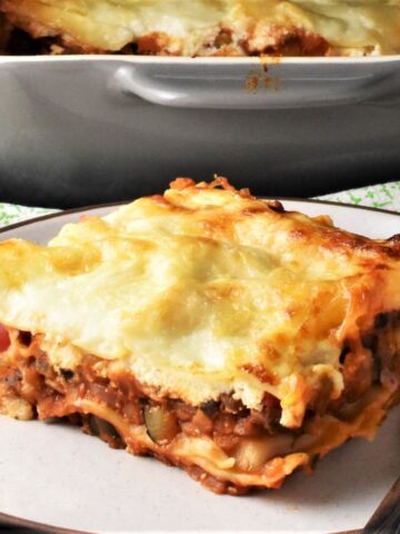 Side view of lentil lasagna piece on white plate with grey casserole dish in background.