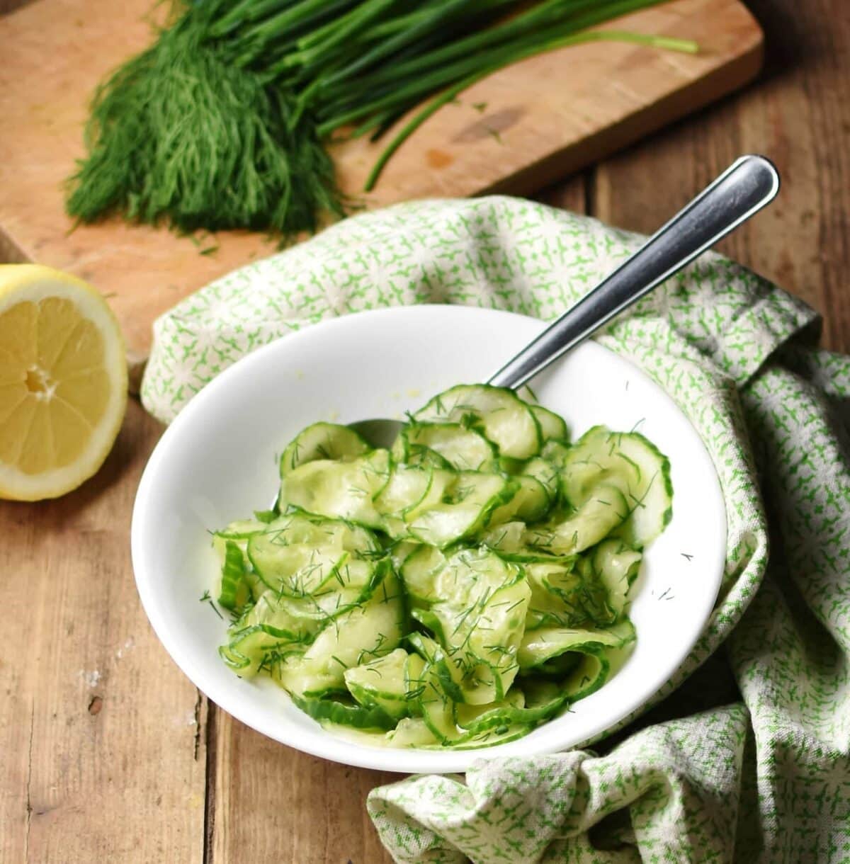Cucumber salad with spoon in white bowl wrapped in green cloth with lemon and herbs in background.