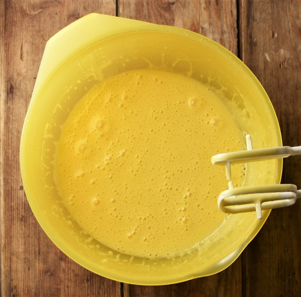 Egg and oil mixture in yellow bowl with electric mixer attachments to the right.