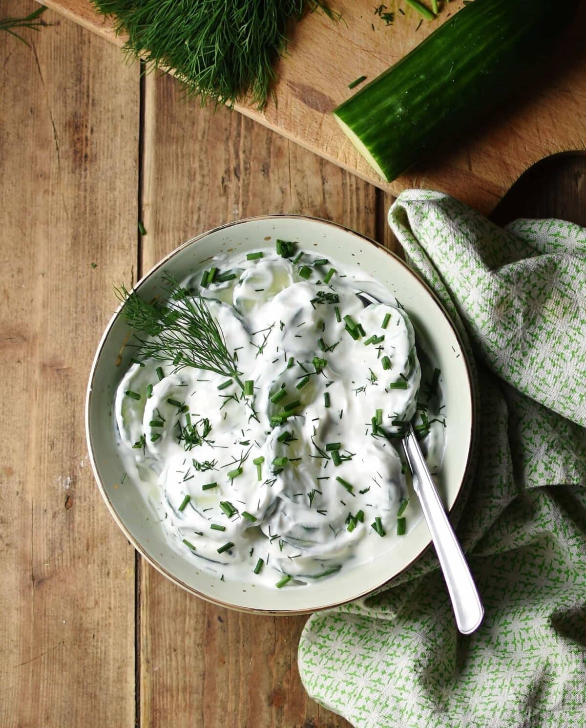 Top down view of creamy cucumber salad with spoon in green bowl wrapped in green cloth, with cucumber and herbs in background.