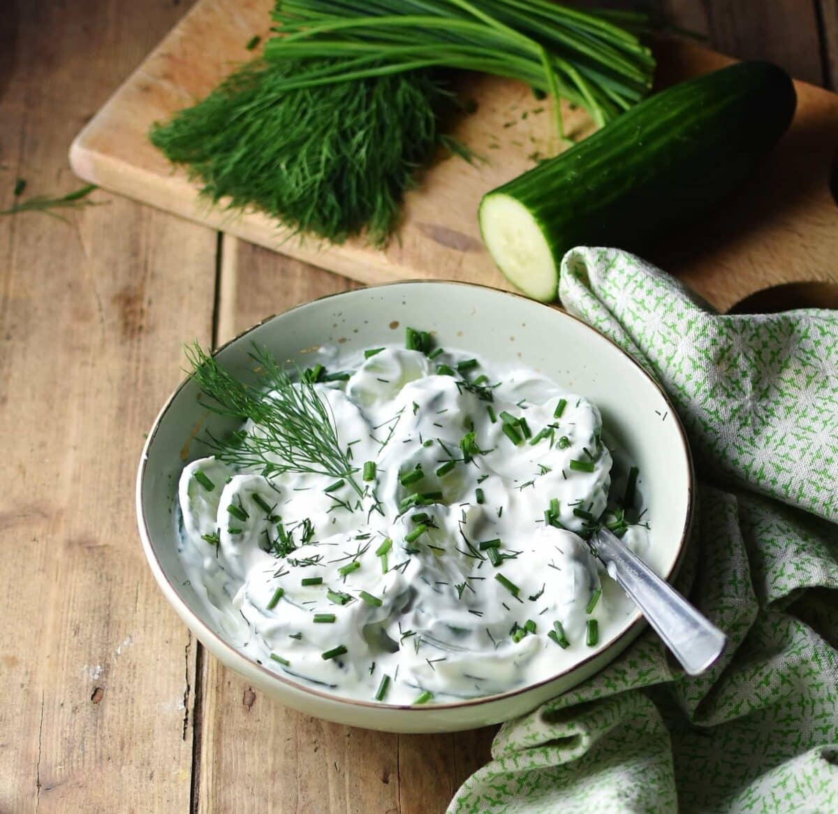 Creamy Polish cucumber salad in green bowl with spoon wrapped in green cloth, with herbs and cucumber in background.
