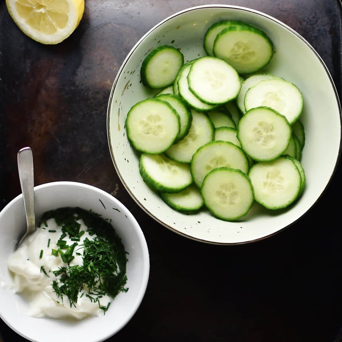 Top down view of creamy sauce in white bowl and cucumber slices in light green bowl.