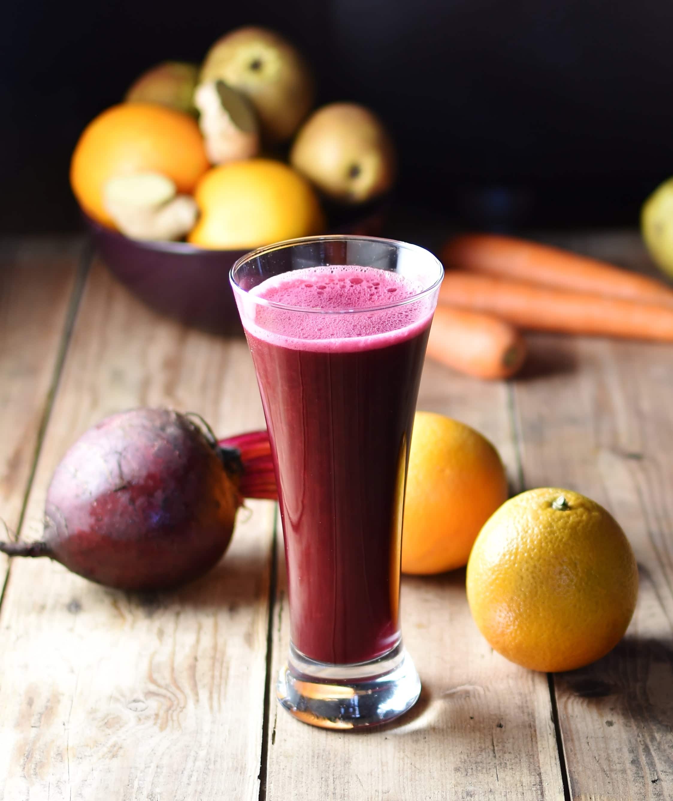 Side view of red cabbage juice in tall glass with 2 oranges, beet and other fruits and vegetables in background.