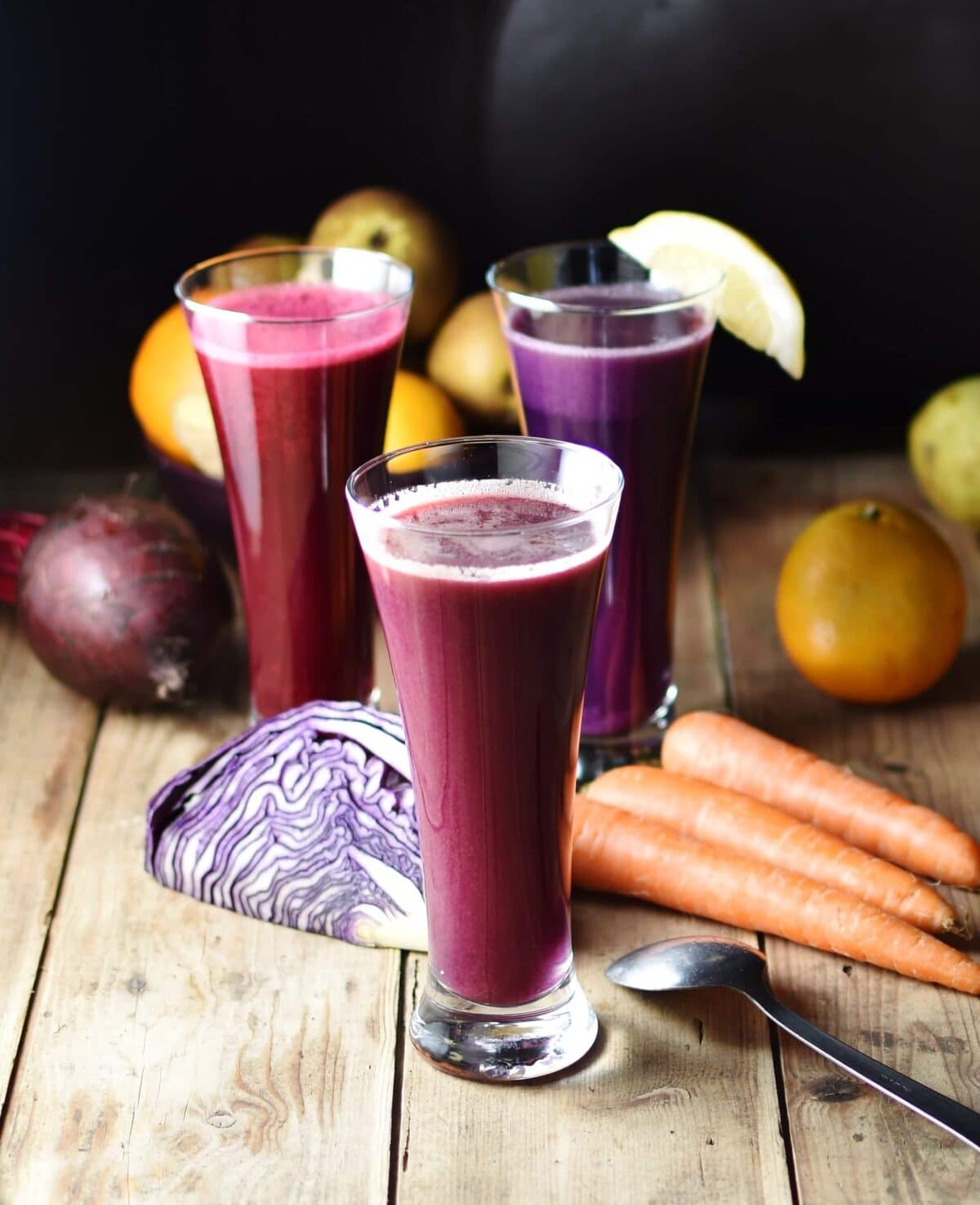 Side view of cabbage juice in 3 separate tall glasses with red cabbage, carrots, beet, and other raw vegetables and fruits in background on top of wooden board with spoon to the right.