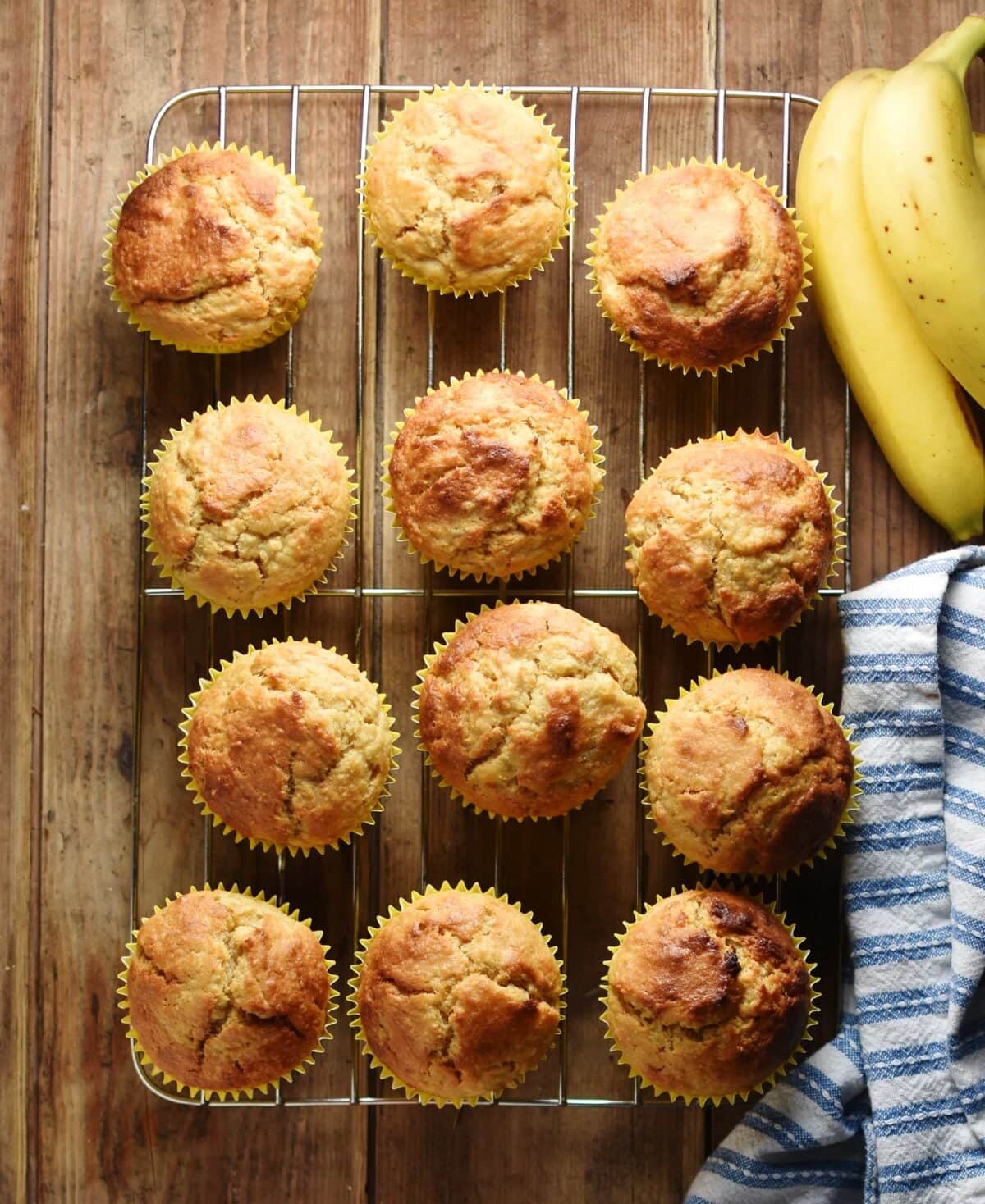 Top down view of banana muffins on top of cooling rack with bananas in top right corner and stripy blue-and-white cloth to the right.