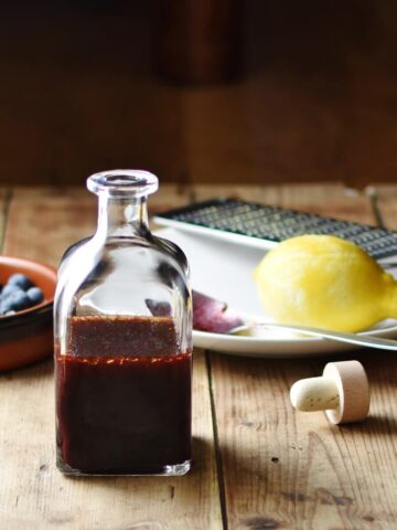 Blueberry dressing in bottle, with blueberries in brown dish, lemon, zester and spoon on top of white plate and bottle top in background.