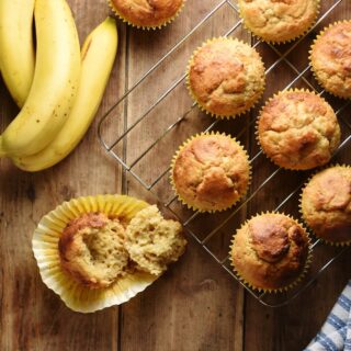 Top down view of banana muffins in yellow cases on top of cooling rack and bananas in top left corner.