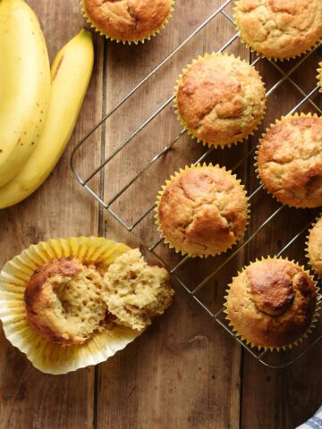 Top down view of banana muffins in yellow cases on top of cooling rack and bananas in top left corner.