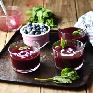Side view of fruit soup in 3 glasses with fresh mint and blueberries in pink cup on top of tray,with mint, blue cloth and dish with spoon in background.