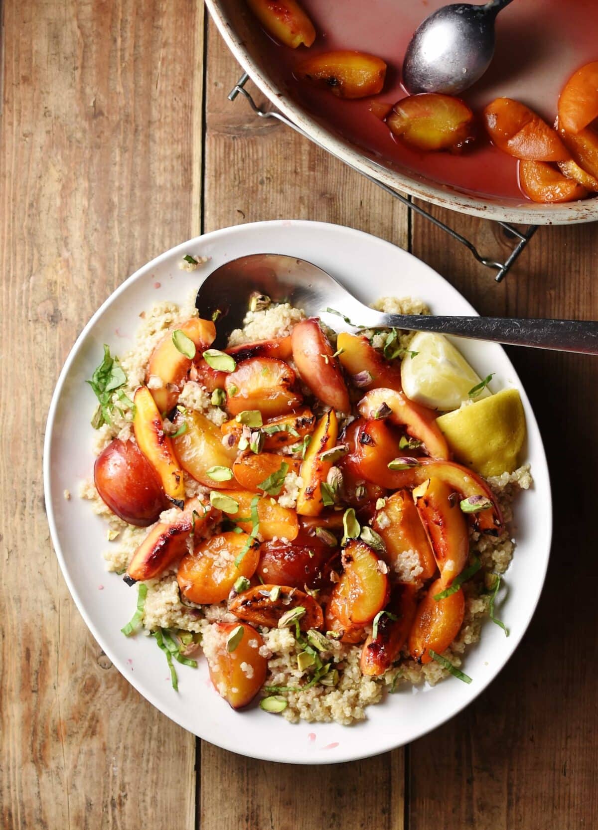 Roasted peach wedges with nuts and herbs on top of quinoa, with lemon wedges and spoon on white plate, with cooked peaches and syrup inside oven dish in top right corner.